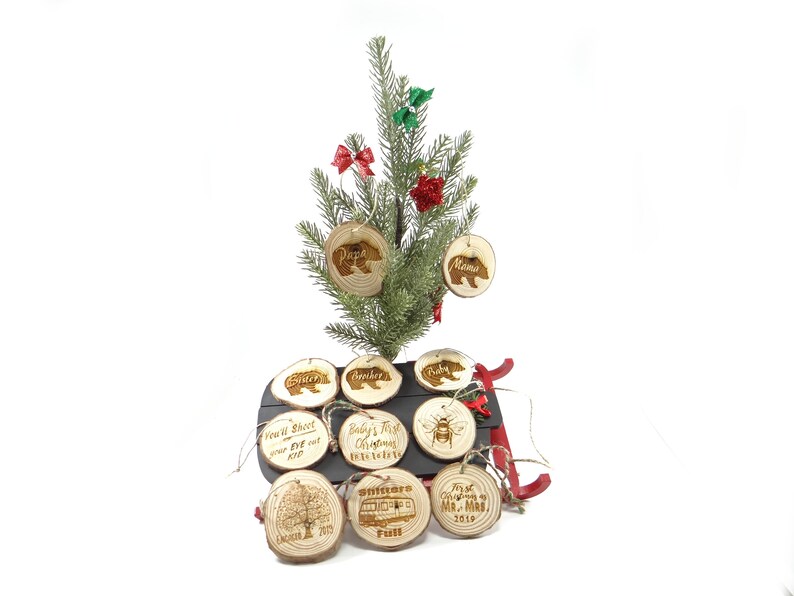 May include: A set of 9 wood slice ornaments with various designs, including a bear, a bee, a tree, a house, and text. The ornaments are hanging on a small artificial Christmas tree and a red sled.