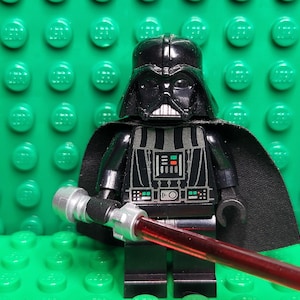 May include: A black Lego Darth Vader figure with a black cape and a red lightsaber. The figure is set against a green Lego baseplate. The figure has a detailed chest plate and helmet.