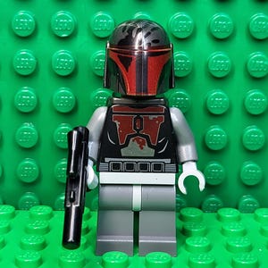 May include: A Lego minifigure of a character with a red and black helmet and armor, holding a black blaster gun. The figure has gray arms and legs, and white hands. The background is a green Lego baseplate.