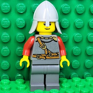 May include: A Lego minifigure of a knight, featuring a yellow head with a smiling face, a white helmet, red arms, gray torso and legs. The torso has a brown harness design. The figure stands on a green Lego base.