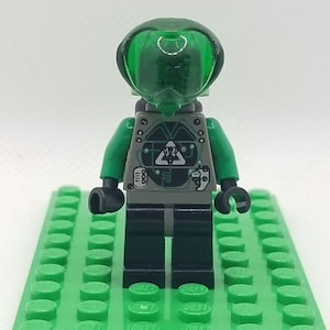May include: A Lego minifigure of an alien with a green transparent helmet and green arms. The torso is gray with a black and white design. The legs are black, and the figure is standing on a green Lego baseplate.