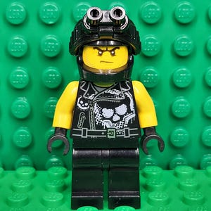 May include: A Lego minifigure with a yellow head and arms, black legs, and a black jacket with a skull design. The figure wears a black helmet with two silver lights. The background is a green Lego baseplate.