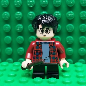 May include: A LEGO minifigure of Harry Potter wearing a red and black plaid shirt, blue sweater, and black pants. He is holding a brown wand in his right hand.