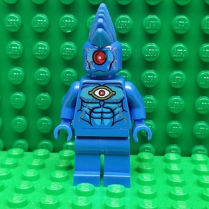 May include: Blue Lego minifigure with a spiky head and a single large eye on its chest. The minifigure has a red eye and a yellow eye on its chest.