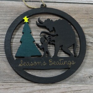May include: Black wooden Christmas ornament with a silhouette of a monster and a child standing next to a green Christmas tree. The ornament has the text "Season's Beatings" written on it.