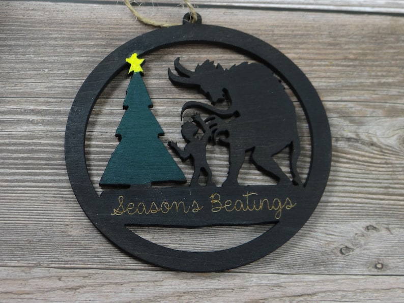 May include: Black wooden Christmas ornament with a silhouette of a monster and a child standing next to a green Christmas tree. The ornament has the text "Season's Beatings" written on it.
