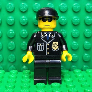 May include: A Lego minifigure of a police officer, wearing a black uniform with a badge and a black hat. The figure has yellow hands and a yellow head with sunglasses. The background is a green Lego baseplate.
