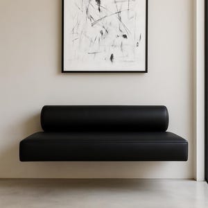 Könnte beinhalten: Ein modernes schwarzes Sofa mit zylindrischer Rückenlehne, an einer Wand montiert. Über dem Sofa hängt ein gerahmtes abstraktes Schwarz-Weiß-Kunstwerk. Der Raum hat ein minimalistisches Design mit neutralfarbenen Wänden und Böden.