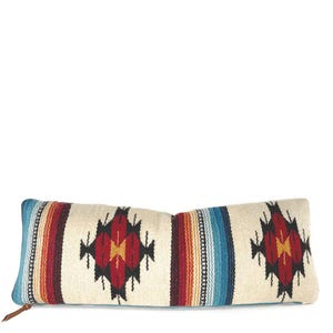 Puede incluir: Una almohada larga y rectangular con fondo blanco y un patrón geométrico colorido. El patrón presenta rayas y diamantes rojos, naranjas, amarillos, azules y negros. La almohada tiene una cremallera de cuero marrón.