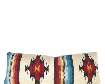 May include: A long, rectangular pillow with a white background and a colorful geometric pattern. The pattern features red, orange, yellow, blue, and black stripes and diamonds. The pillow has a brown leather zipper.
