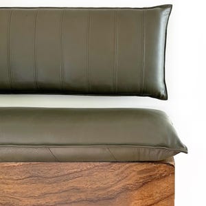 May include: A dark green leather bench cushion with a wooden base. The cushion is made of two pieces, one for the seat and one for the back. The back cushion has vertical stitching.