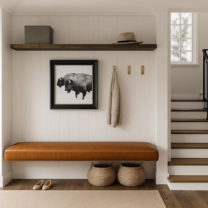 Könnte beinhalten: Ein Hauseingang mit einer braunen Lederbank, einem gerahmten Schwarz-Weiß-Bison-Druck und einem Holzregal mit Hut und Box. Zwei geflochtene Körbe stehen unter der Bank. Ein beiger Mantel hängt an Messinghaken.