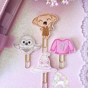May include: Four decorative paper clips featuring embroidered designs. Includes a light brown elf, a white owl, a pink sweater, and a white witch hat with pink accents. The clips are on a pink glittery surface.
