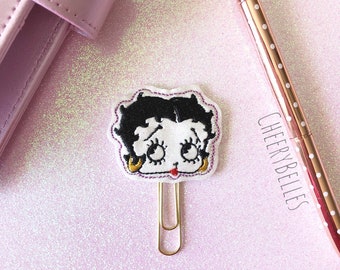 May include: A Betty Boop-themed paper clip with a gold finish, featuring a white face, black hair, and a red lip. The clip is on a pink glitter surface, next to a rose gold pen with a diamond-shaped top and the text "CHEERYBELLES".