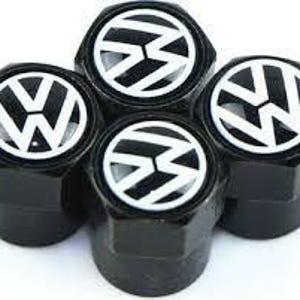 May include: Four black tyre valve stem caps with a white Volkswagen logo. The caps are hexagonal and have a glossy finish. The Volkswagen logo is a stylized "VW" emblem.