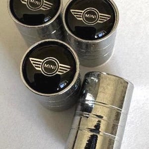 May include: A set of four cylindrical tyre valve stem caps with a polished chrome finish. Each cap features a circular black inlay on the top, displaying the white MINI logo with stylised wings and the word MINI centred within the circle.