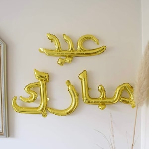 Könnte beinhalten: Goldene Folienballons, die "Eid Mubarak" in arabischer Schrift buchstabieren. Die Ballons hängen an einer weißen Wand.