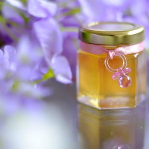Könnte beinhalten: Ein kleines Glas gefüllt mit Honig, gebunden mit einem rosa Band und einem rosa Schnuller-Anhänger. Das Glas steht auf einem Bett aus lila Blüten.