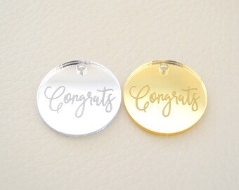 May include: Two round, mirrored acrylic tags with the word "Congrats" engraved on them. One tag is silver and the other is gold.