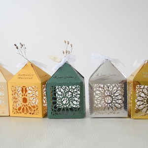 Puede incluir: Cinco cajas de regalo decorativas para el Ramadán en diferentes colores, incluyendo crema, amarillo, verde, plata y oro. Cada caja presenta un diseño delicado e intrincado cortado con láser y las palabras "Ramadán Mubarak" en escritura árabe.