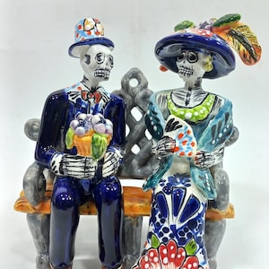 Puede incluir: Una figurita de cerámica de una pareja vestida con atuendos tradicionales mexicanos del Día de Muertos, sentada en un banco. El hombre lleva un traje azul y un sombrero de copa, mientras que la mujer lleva un vestido azul y verde con un gran sombrero. Ambos llevan flores.