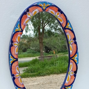 Puede incluir: Espejo ovalado con un marco de cerámica pintado a mano y colorido. El marco presenta un diseño floral azul, naranja y blanco.