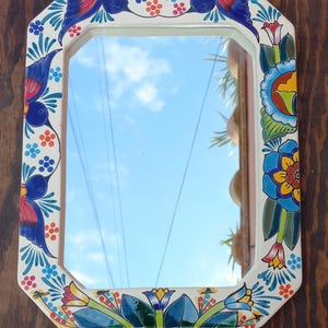 May include: An octagonal mirror with a white frame, adorned with colorful floral and bird designs. The mirror reflects a blue sky with clouds. The frame features blue, red, yellow, and green accents, creating a vibrant, decorative piece.