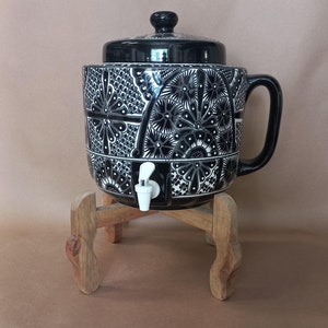 May include: Black and white ceramic water dispenser with a wooden stand. The dispenser has a floral design and a spigot.