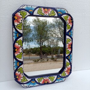 May include: A decorative mirror with a blue, white, and green floral design. The mirror is framed in a blue and white ceramic tile with a floral pattern. The mirror reflects a view of trees and a road.