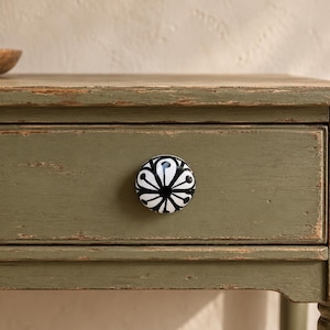 May include: A round ceramic drawer knob featuring a black and white floral pattern with radiating petals. The knob is attached to a rectangular wooden drawer front finished in a distressed olive green paint with visible wood grain texture.