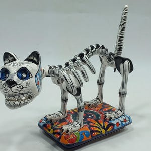 May include: A Day of the Dead cat figurine. The ceramic cat has a white body with black stripes and a skeleton design. The head is white with blue eyes and floral accents. It rests on a colorful, patterned base.