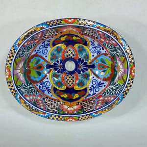 Op de afbeelding: Een ovale keramische wastafel voor de badkamer met een levendig, handgeschilderd patroon in Talavera-stijl. Het ontwerp bevat ingewikkelde bloemmotieven en geometrische vormen in tinten kobaltblauw, feloranje, geel, groen en roze.