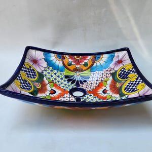 May include: A rectangular ceramic vessel sink with curved sides and a dark blue rim. The interior features a vibrant, multi-colored hand-painted floral and geometric pattern in shades of red, yellow, light blue, green, and white, with a central drain hole.