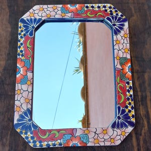 May include: Rectangular mirror with a colorful floral design. The frame features a repeating pattern of orange, blue, and white flowers with green accents. The mirror reflects a blue sky and a building.