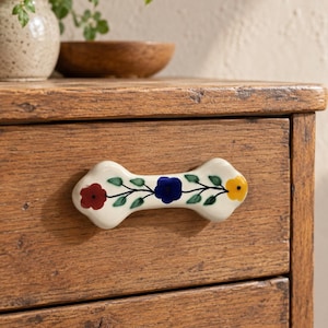 May include: A cream-colored ceramic drawer pull with a hand-painted floral motif featuring a red flower, a blue flower, and a yellow flower connected by green vines and leaves, mounted horizontally on a rustic wooden drawer front.