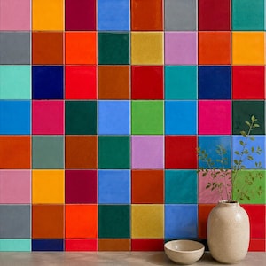 May include: A wall covered in a grid of square, glossy ceramic tiles in a vibrant array of colors including red, blue, green, yellow, orange, pink, and purple. A beige ceramic vase containing a leafy branch and a small beige bowl sit in front.