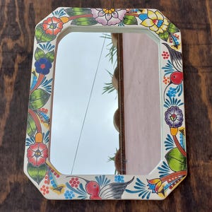 May include: Rectangular decorative mirror with a cream-colored frame. The frame is adorned with colorful floral and bird designs in red, blue, yellow, and green. The mirror reflects the sky and surrounding environment.