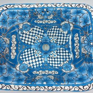 Pode incluir: Uma pia quadrada de cerâmica com um design floral azul e branco. A pia tem um furo de drenagem no centro e uma borda branca.