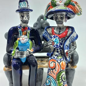 Puede incluir: Figura de cerámica de una pareja del Día de Muertos, sentada en un banco gris. La figura masculina viste un traje y sombrero azules, sosteniendo un ramo floral azul y verde. La figura femenina viste un vestido y sombrero coloridos con detalles florales.