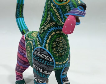 May include: A vibrantly painted wooden alebrije sculpture of a dog-like creature with a long tail and large ears. The folk art figure features intricate patterns in green, blue, pink, and gray, with a pink tongue extending from its mouth.