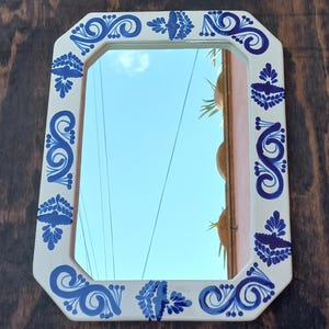 May include: An octagonal mirror with a cream-colored frame adorned with blue floral patterns. The mirror reflects a clear blue sky and is set against a dark wood background. The frame's design adds a decorative touch.
