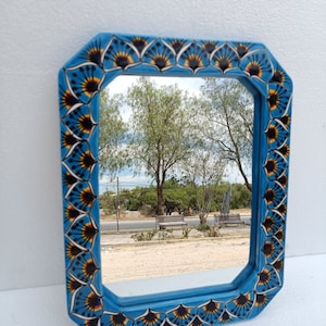 May include: A square, blue-framed mirror with a decorative border featuring a repeating pattern of black, yellow, and blue fan-like shapes. The mirror reflects an outdoor scene with trees and benches.