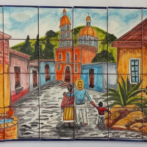 May include: A vibrant ceramic tile mural showing a village scene. The artwork features a family walking towards a church with orange domes. Buildings in shades of orange, pink, and blue line the cobblestone street. A well and cacti are also visible.