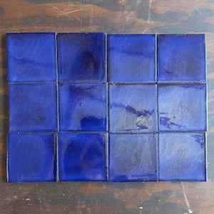 May include: A collection of sixteen square, cobalt blue tiles arranged in a grid pattern. The tiles have a glossy finish, reflecting light and creating subtle variations in color. The tiles are likely for use in a mosaic or as decorative elements.