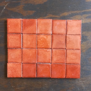 May include: A collection of twenty-four square, terracotta-colored tiles arranged in a rectangular grid. The tiles have a glossy finish and vary slightly in shade, creating a textured appearance. The tiles are set against a dark wood surface.