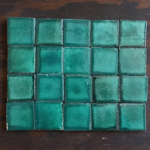 May include: A collection of twenty-four square, emerald green ceramic tiles arranged in a rectangular grid. The tiles have a glossy finish and a slightly uneven surface, creating a textured appearance. The tiles are displayed on a dark brown wooden surface.