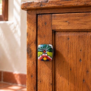 May include: A square ceramic cabinet knob featuring a hand-painted floral pattern in vibrant shades of green, yellow, red, and blue, mounted on a rustic, weathered brown wooden cabinet door with visible wood grain and texture.