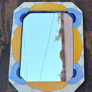 May include: Decorative rectangular mirror with a blue, yellow, and white painted ceramic frame. The mirror's frame features a repeating pattern of stylized floral or leaf-like shapes. The mirror reflects a blue sky and a glimpse of a building.