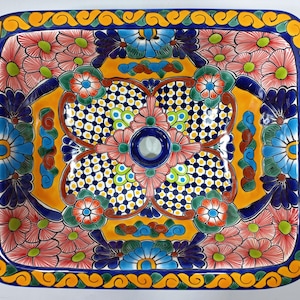 Puede incluir: Lavabo rectangular de cerámica con un vibrante diseño floral. El lavabo presenta un centro amarillo con un diseño de cuadros azules y blancos, rodeado de flores rosas, azules y naranjas. El borde es azul con un patrón de desplazamiento amarillo.