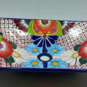May include: Rectangular ceramic sink with a vibrant floral design. The sink has a deep blue border and is decorated with colorful flowers in shades of red, blue, yellow, and green. The background has a white and brown lattice pattern.